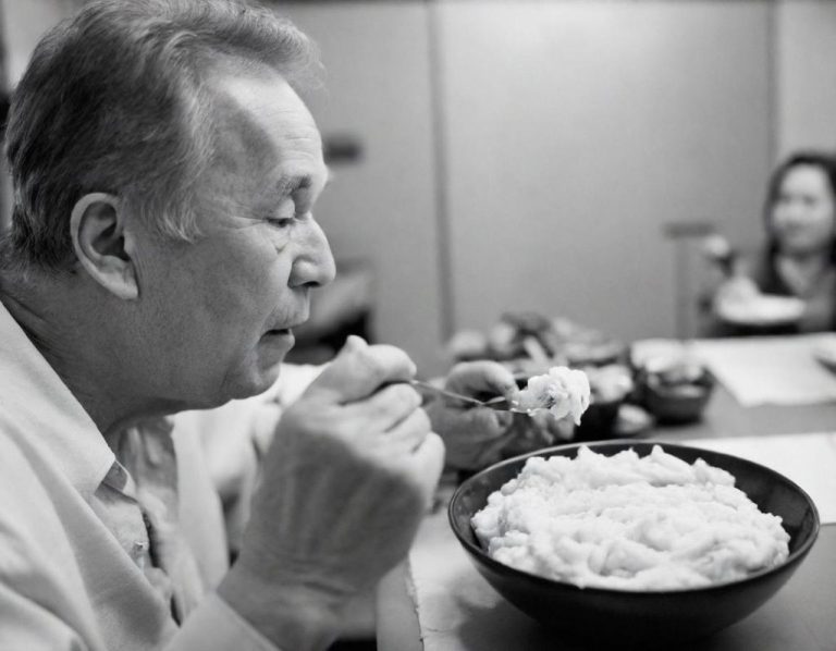 Plat en texture modifié dans une assiette creuse sans présentation particulière Homme âgé savourant un grand bol de purée, assis à une table avec une femme en arrière-plan.